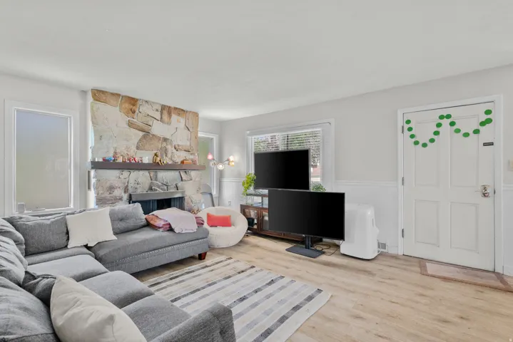 Spacious living area featuring a stone-clad fireplace with a wood mantle, light wood-finish flooring, white walls with wainscoting, and large windows