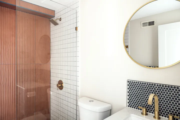 Modern bathroom featuring a fluted glass shower enclosure with brass fixtures, a white subway tile shower surround, a circular mirror with a brass frame, a sink with a brass faucet, and a black penny tile backsplash