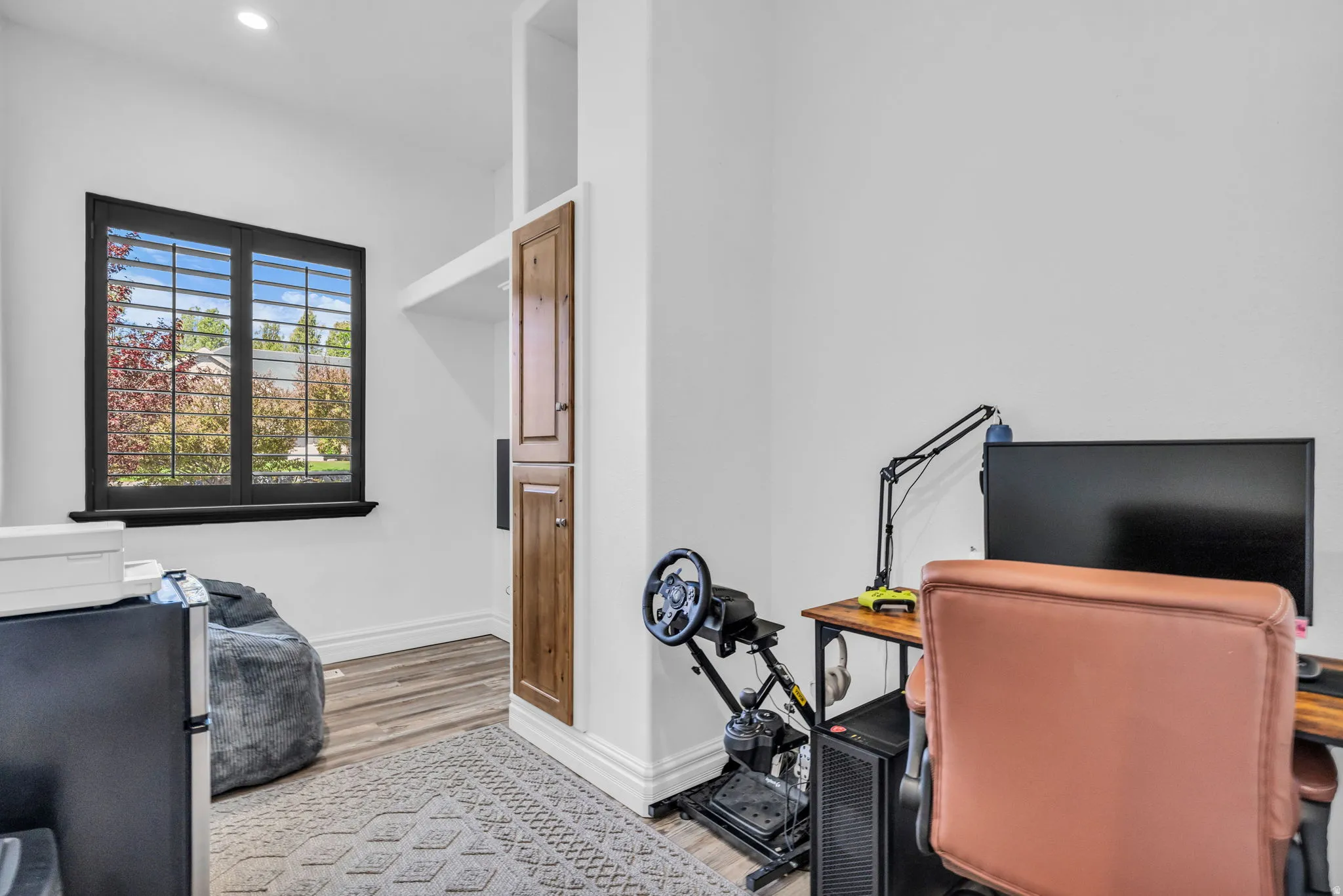Bright room featuring a black-framed window with louvered shutters, recessed ceiling lighting, built-in wood-finish cabinetry, and wood-finish flooring