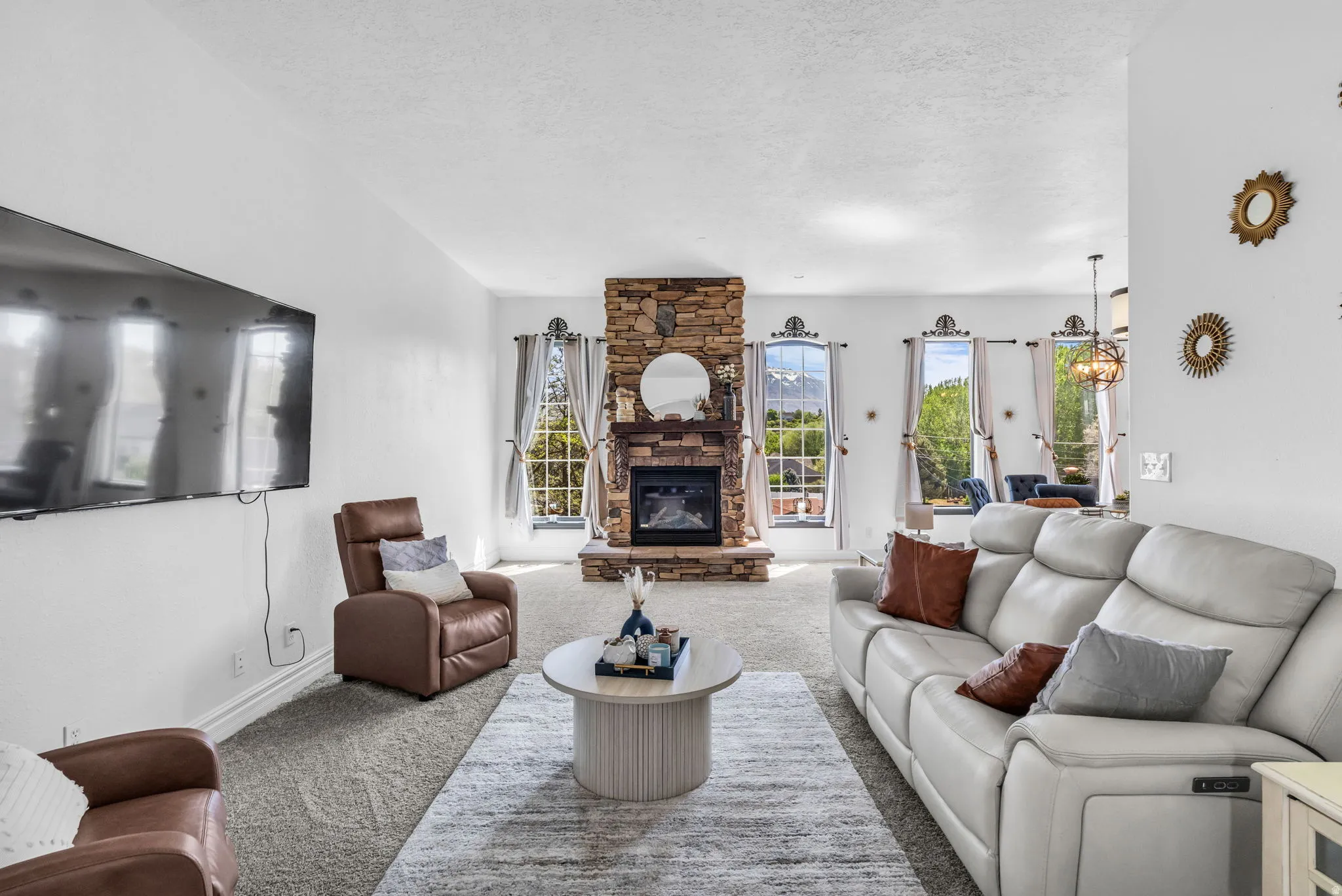 Stone-faced fireplace with hearth, multiple windows, integrated wall-mounted television, and neutral carpeting