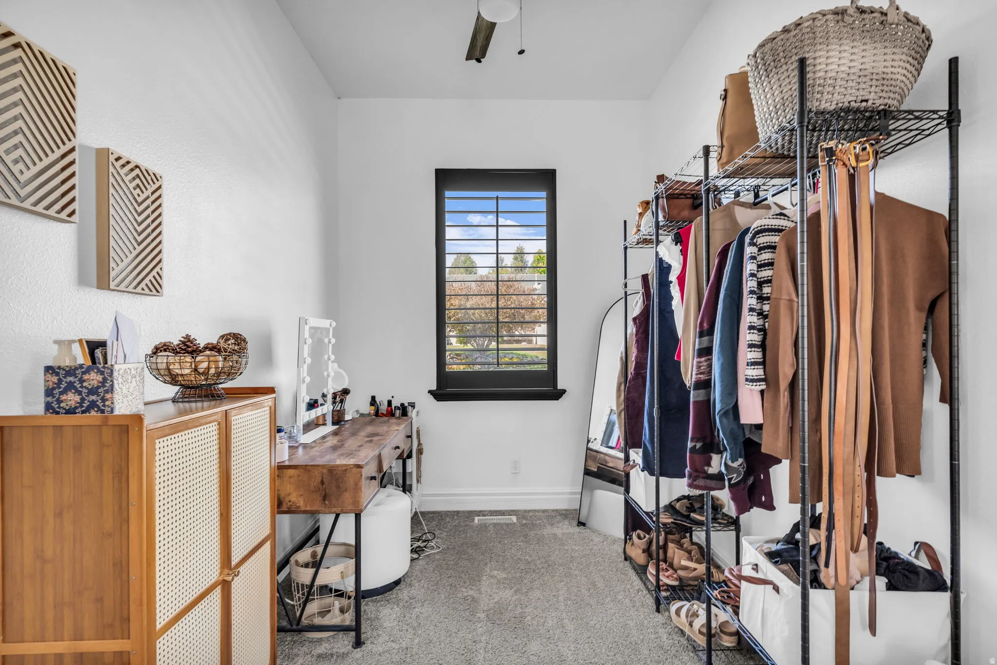 Carpeted dressing area featuring a window with dark plantation shutters, a wood-finish vanity, and a ceiling fan