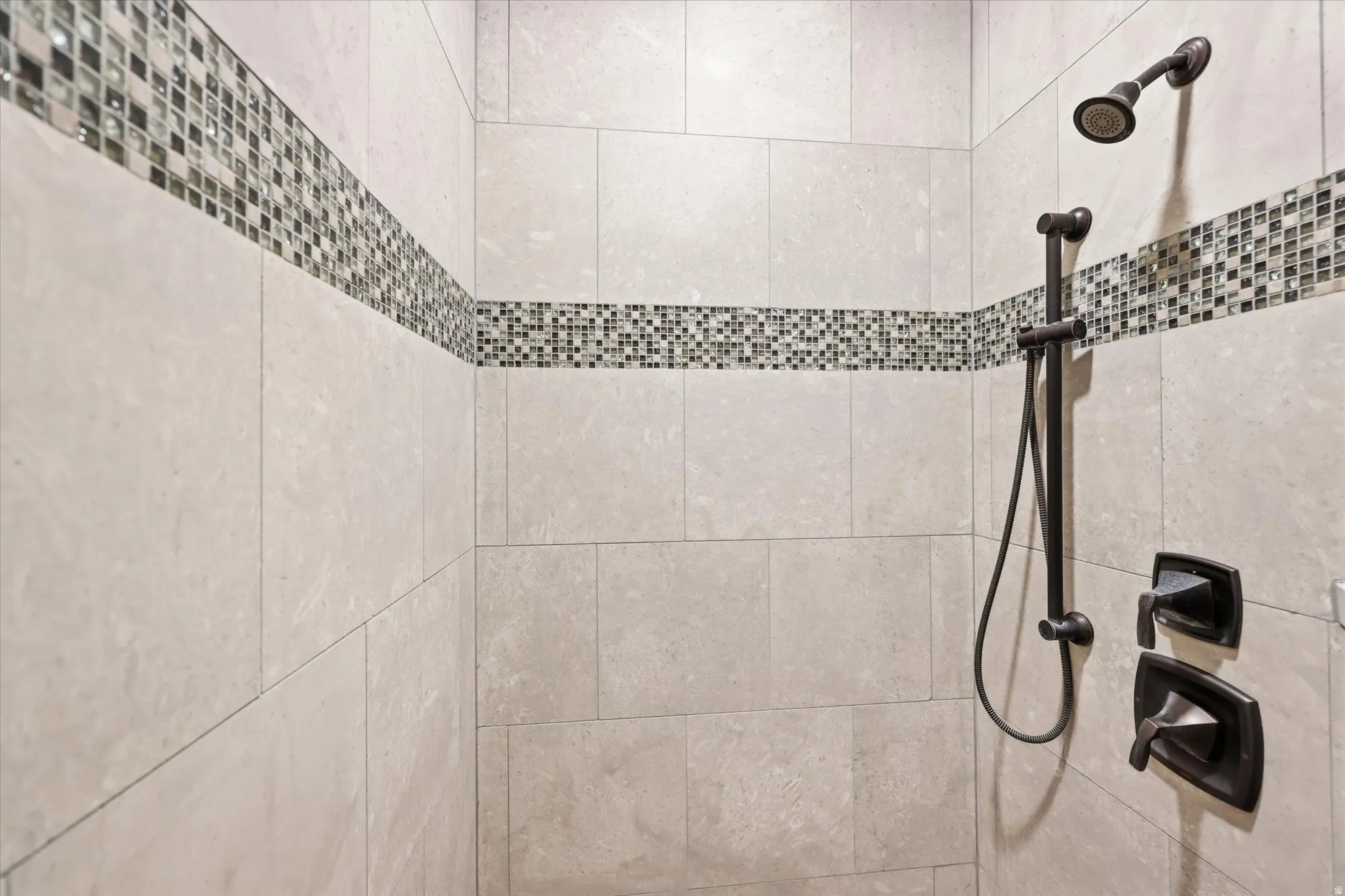 PRIMARY Shower enclosure featuring large format stone-look tiles, a mosaic tile accent band, a fixed shower head, and a handheld shower attachment with bronze finishes