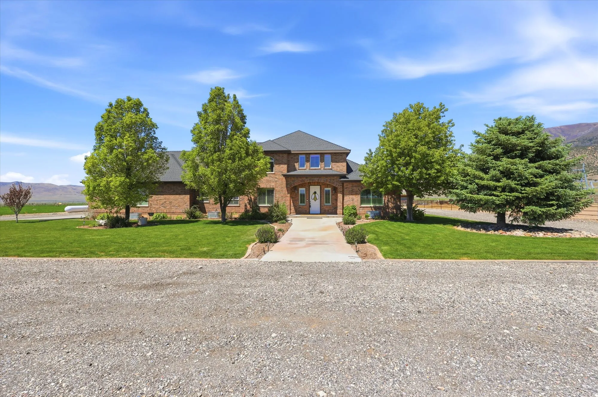 Brick facade residence featuring a front entry archway, a paved walkway, established trees, a manicured lawn, and a graveled drive