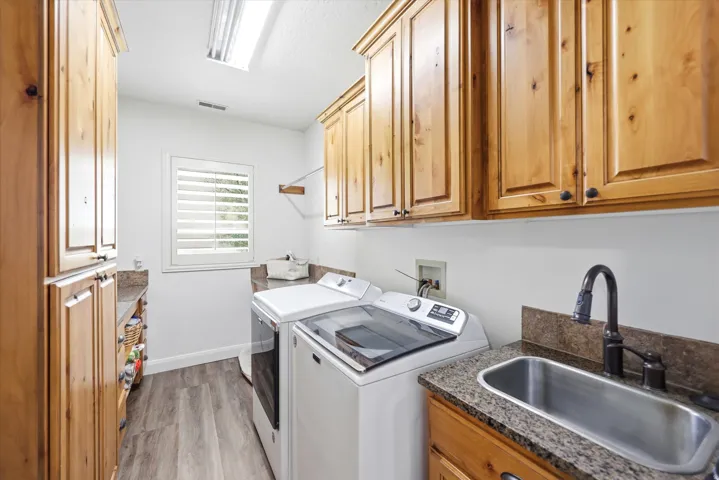 Dedicated utility room featuring wood-finish cabinetry, granite-style countertops, stainless steel sink with a dark-finish faucet, and wood-finish flooring