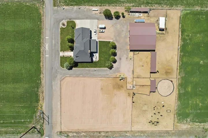 Expansive rural property featuring a main residence with a dark gray roof, a large agricultural building with a maroon roof, multiple fenced enclosures, an open dirt arena, and surrounding green fields