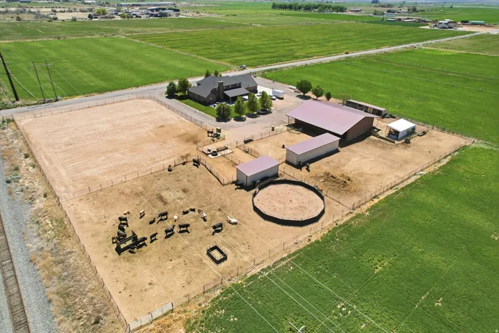 Expansive rural property featuring a brick residence, multiple outbuildings with metal roofs, fenced animal enclosures including a round pen, and a large, uncultivated field