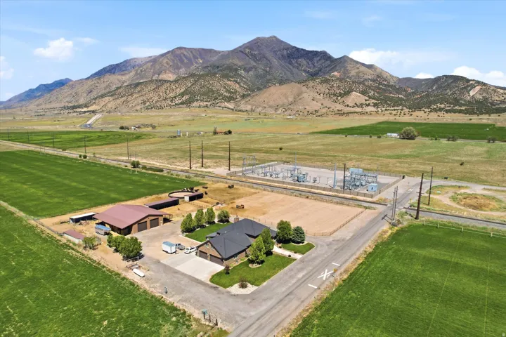 Expansive property featuring a brick residence with an attached garage, a large brown barn with multiple bays, a circular arena, and vast green fields extending towards the mountains