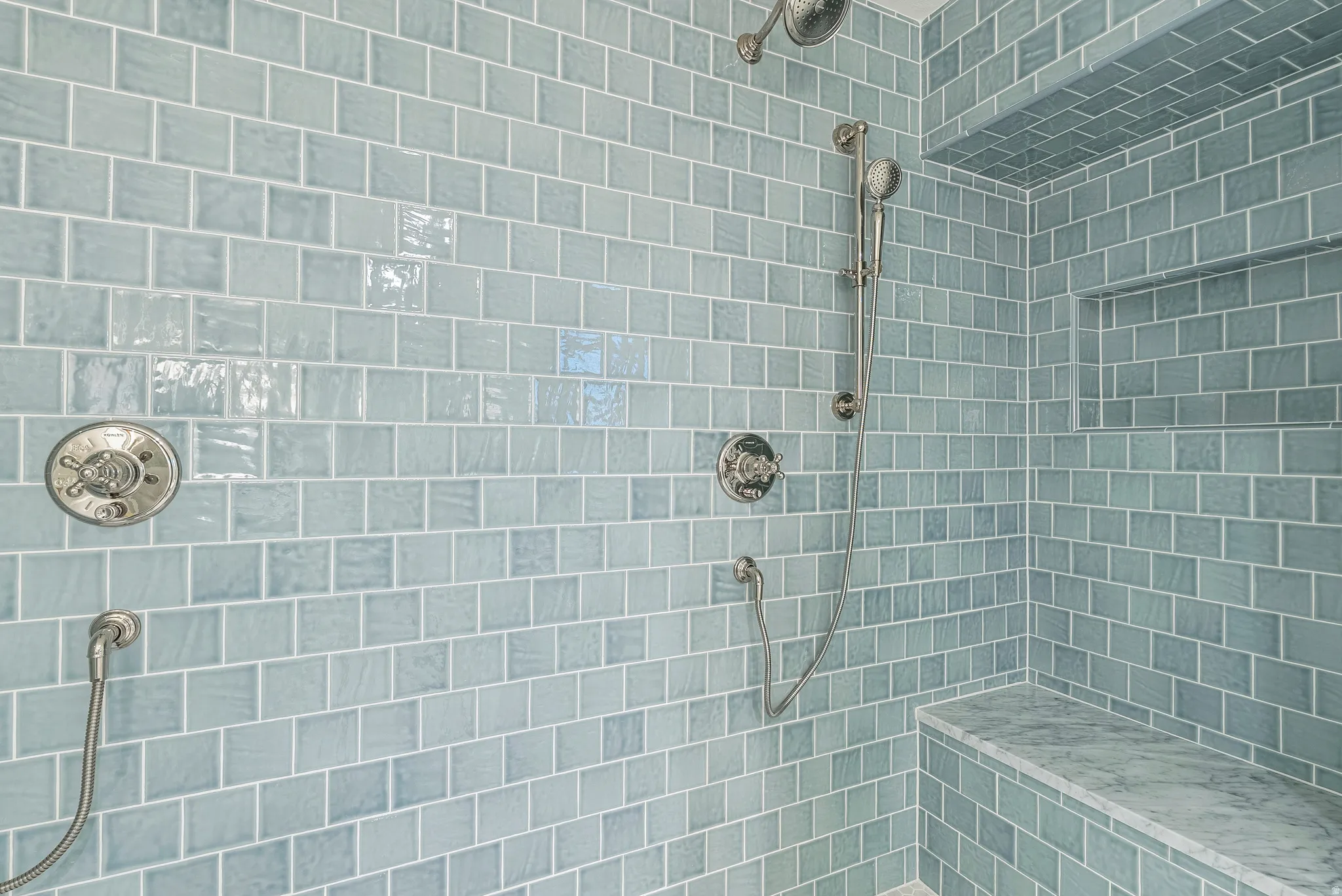 Shower enclosure featuring light blue subway tile, a built-in bench with a stone surface, a recessed niche, a fixed shower head, and a handheld shower attachment