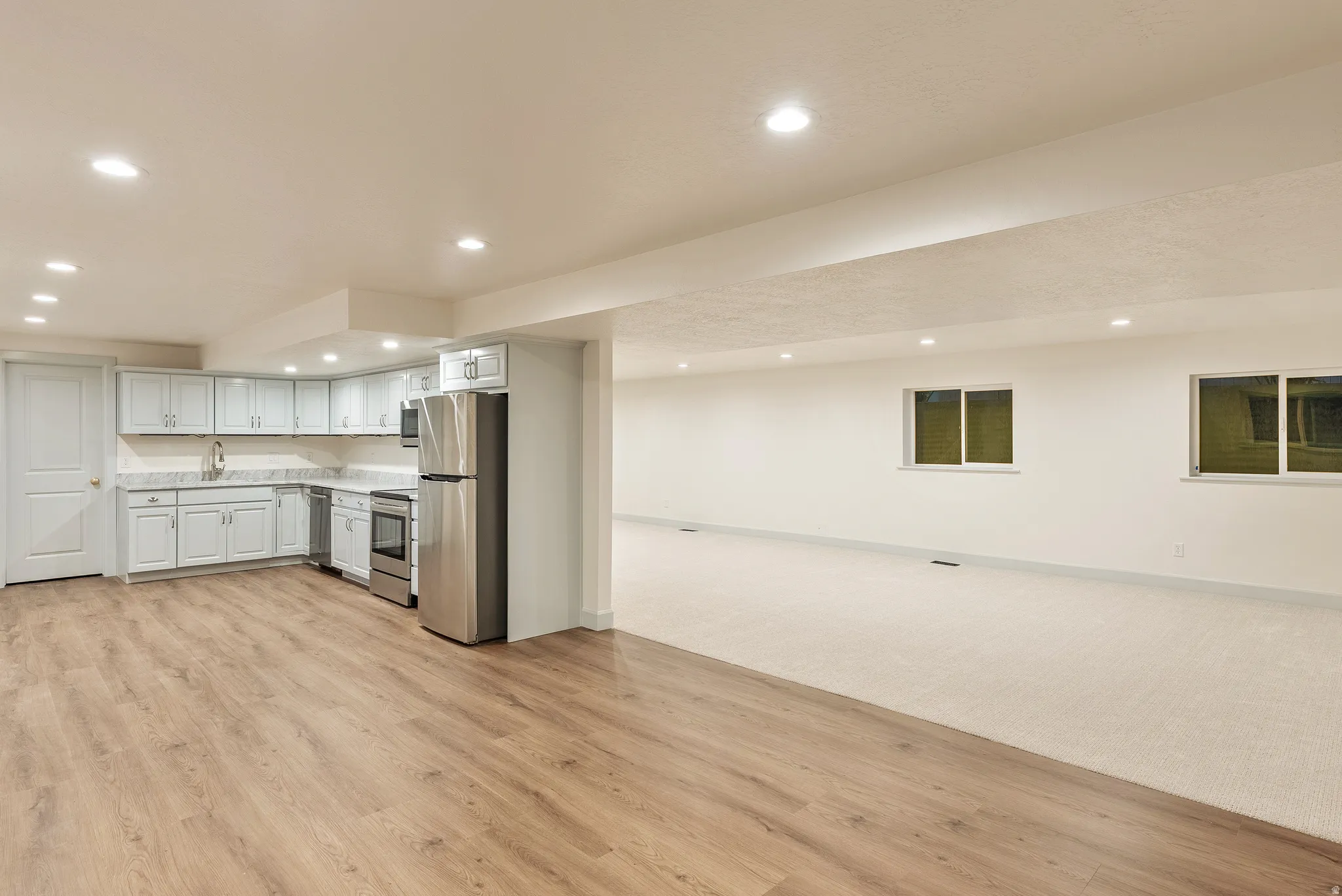Open-concept living area featuring wood-finish flooring, a full kitchen with light cabinetry and stainless steel appliances, recessed lighting, and two windows