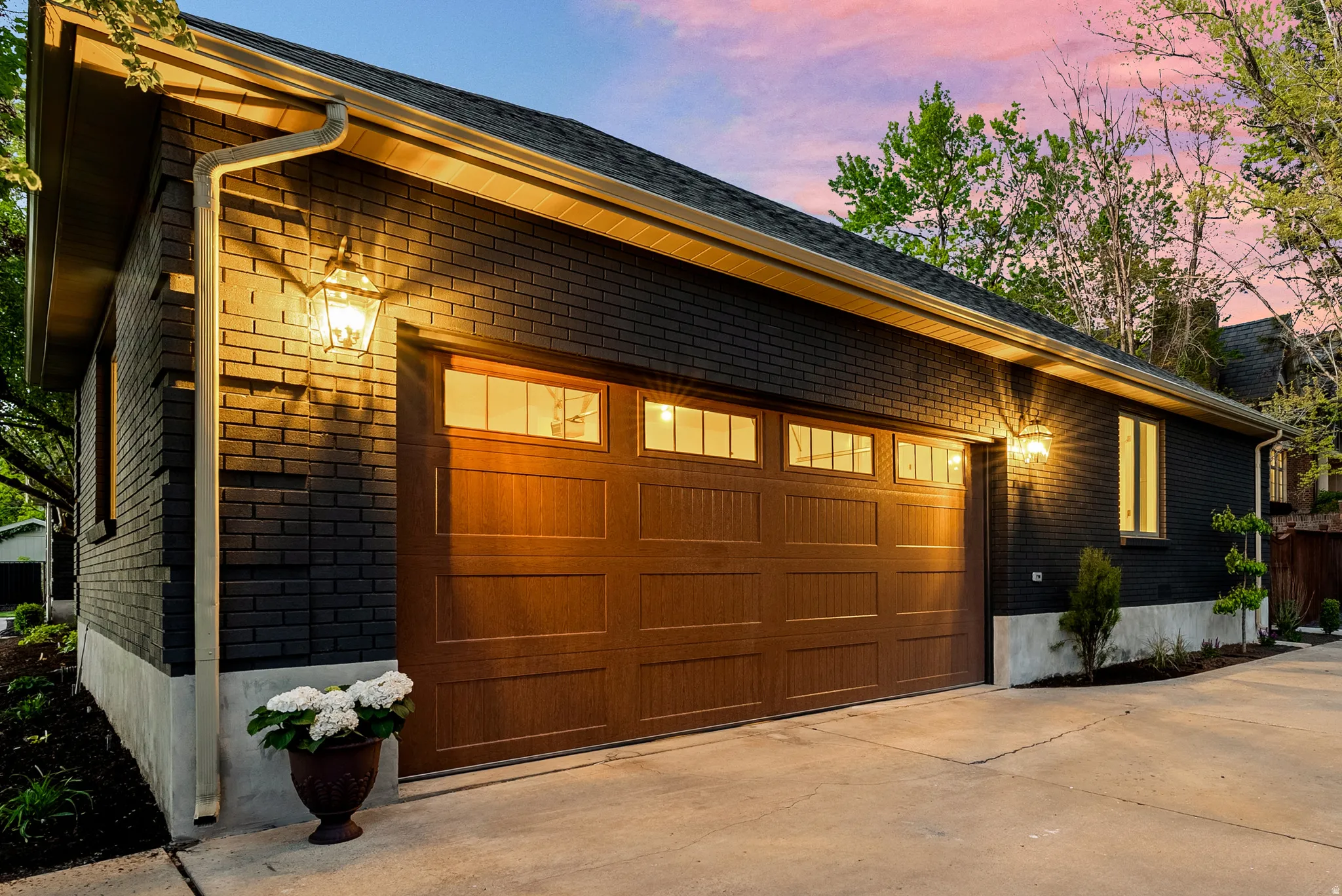 Detached garage featuring dark brick exterior, a wood-finish garage door with upper window inserts, exterior wall lanterns, and a concrete driveway
