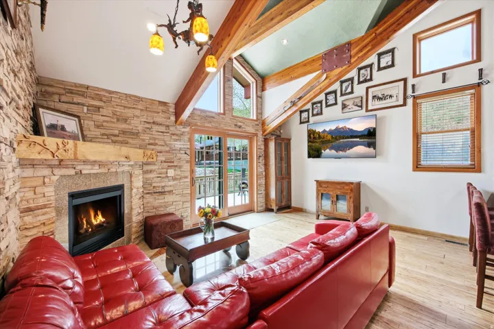 Living area featuring a stone accent wall with a built-in fireplace, vaulted ceilings with exposed wood beams, wood-finish flooring, and sliding glass doors opening to the exterior