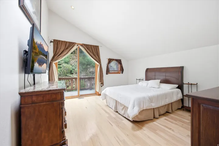Vaulted ceiling bedroom with light wood-finish flooring, featuring sliding glass doors to a private deck, a brown wood-frame window, and recessed lighting
