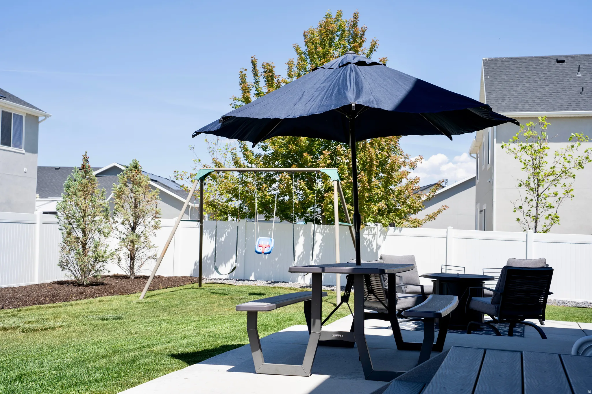Expansive backyard featuring a concrete patio, verdant lawn, mature privacy trees, and a white vinyl privacy fence