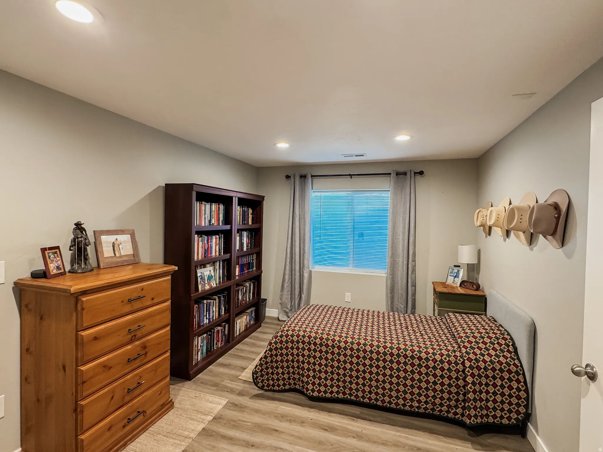 Recessed lighting illuminates this room featuring wood-finish flooring, light gray wall paint, and a window with blinds and gray curtains