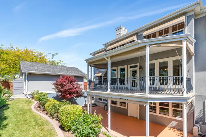 Rear exterior featuring a covered balcony with wrought iron railing, multiple French doors, and retractable awnings