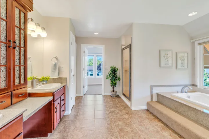Spacious bathroom featuring dual wood-finish vanities with integrated sinks, a tiled shower enclosure, a built-in soaking tub with steps, and neutral floor tiling