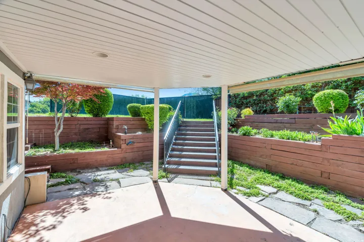 Covered patio with concrete flooring and stone pavers, featuring tiered landscaping with rich wood-toned retaining walls and mature greenery