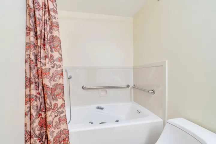 White corner jetted tub with integrated shelving, chrome grab bars, and a wall-mounted showerhead
