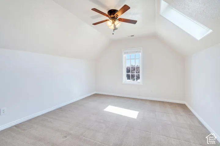 Room featuring a vaulted ceiling, a skylight, a window with exterior views, a ceiling fan with integrated lighting, and carpet flooring