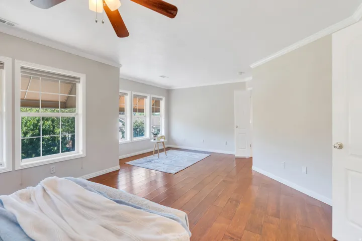 Spacious room featuring rich wood-finish flooring, multiple windows providing ample natural light, bright white trim work, and a ceiling fan with integrated lighting