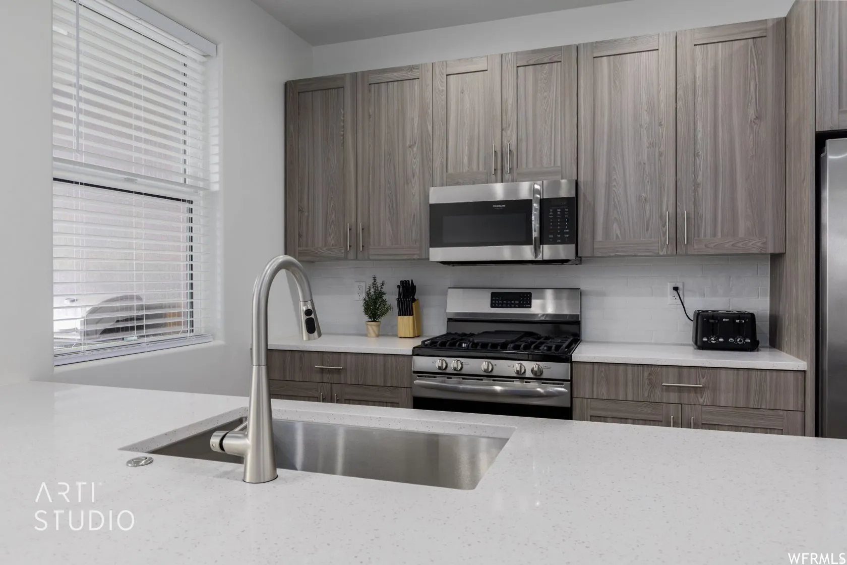 Kitchen featuring light wood-finish cabinetry, stainless steel appliances, a tile backsplash, light stone countertops, and a stainless steel undermount sink with a gooseneck faucet