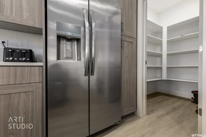 Stainless steel side-by-side refrigerator, wood-finish cabinetry, light-toned countertop, subway tile backsplash, and a spacious walk-in pantry with extensive shelving