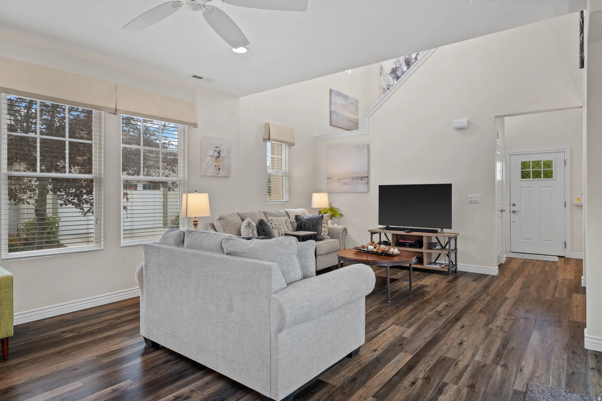 Spacious living area featuring wood-finish flooring, high ceilings, a ceiling fan, and multiple windows with blinds
