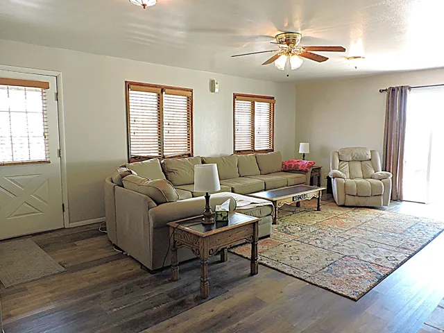 Spacious living area featuring wood-finish flooring, a ceiling fan with integrated lighting, and multiple windows with blinds