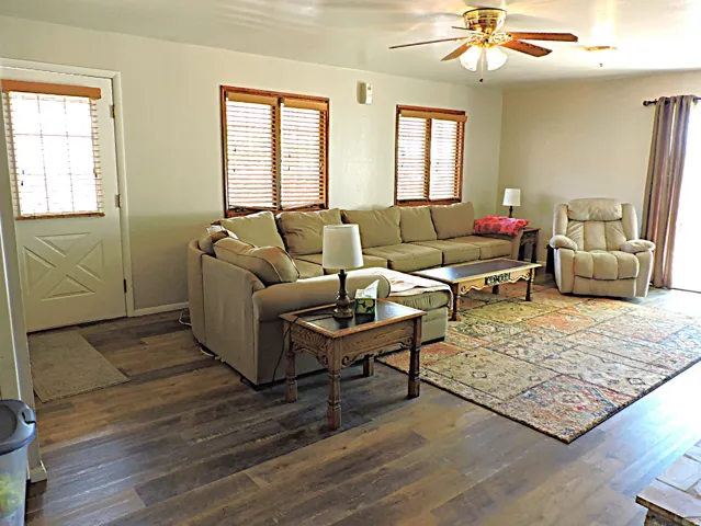 Spacious living area featuring wood-finish flooring, a ceiling fan with integrated lighting, and multiple windows with blinds