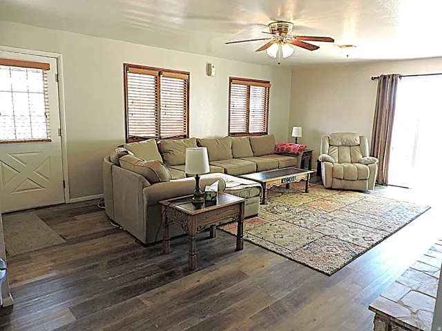 Spacious living area featuring wood-finish flooring, a ceiling fan with integrated lighting, and multiple windows with blinds