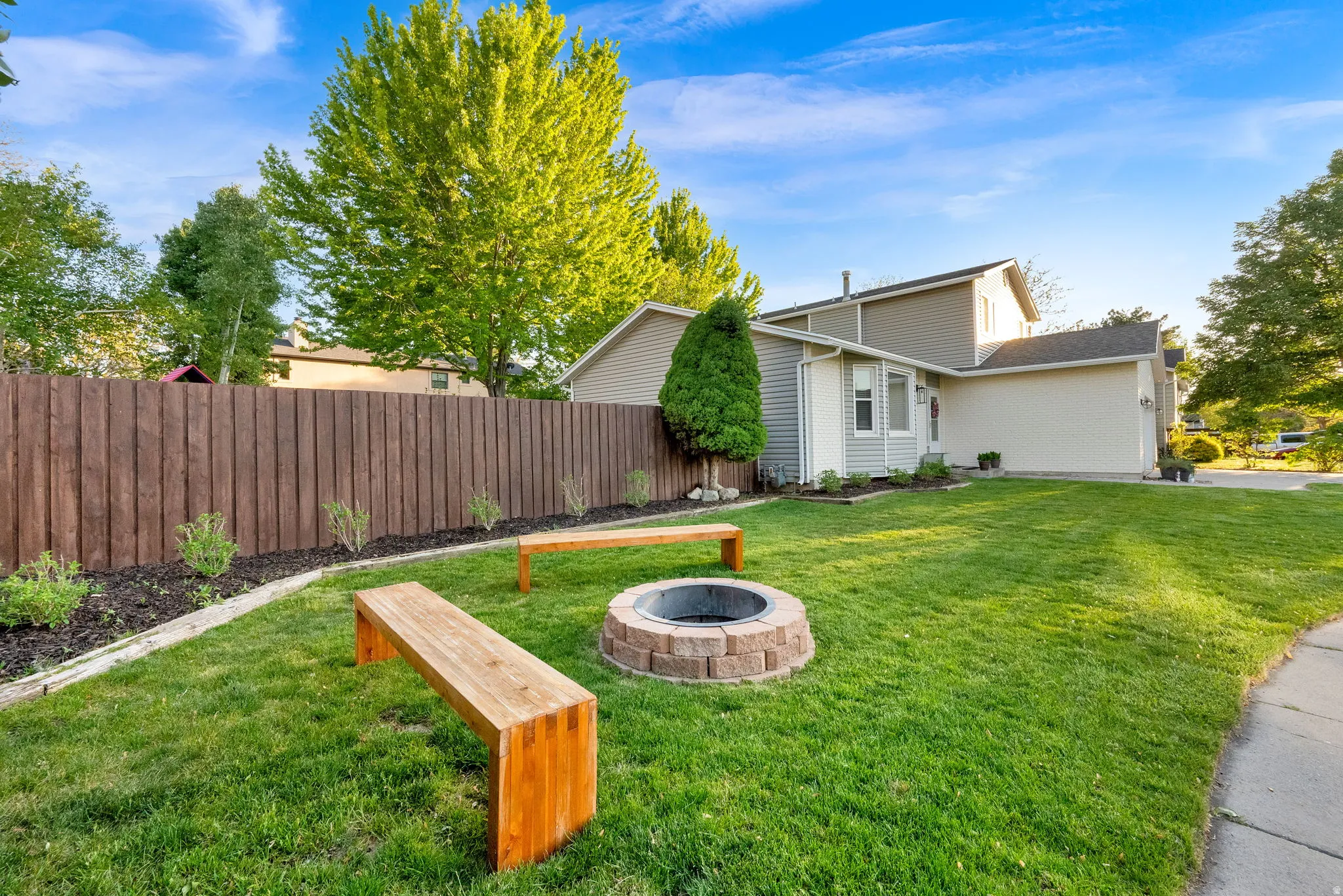 Expansive lawn area featuring a stone fire pit with two wooden benches, a dark wood privacy fence, and mature trees