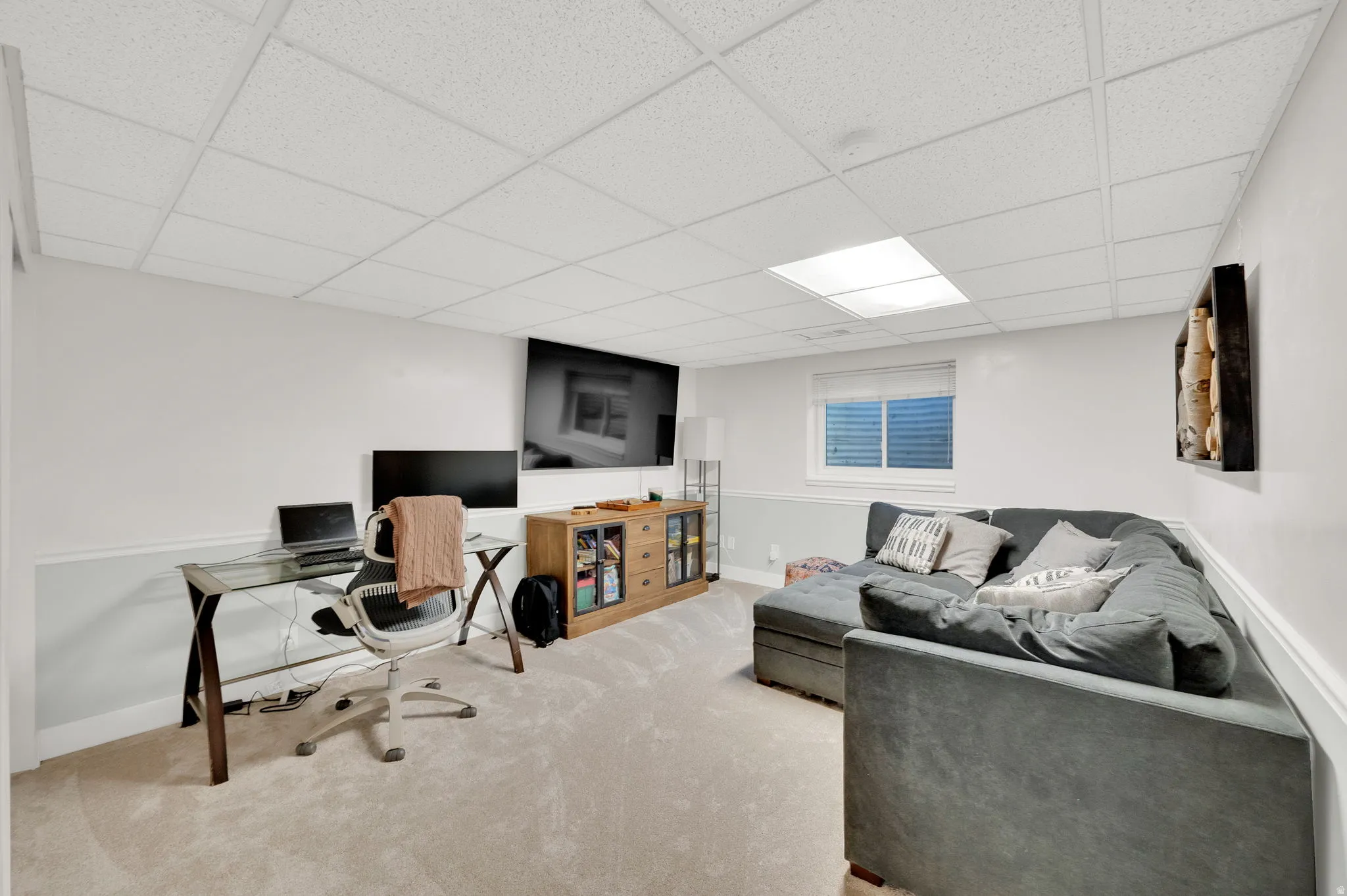 Finished lower level featuring neutral carpeting, white wainscoting, and a drop ceiling with integrated lighting