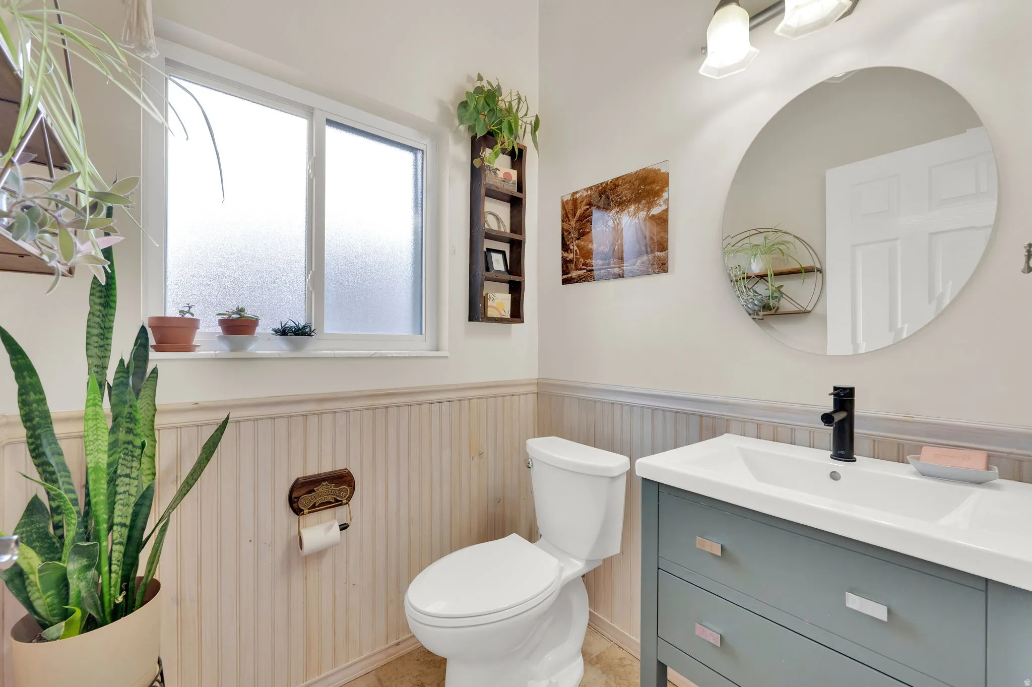 Bathroom featuring a frosted window, beadboard wainscoting, a modern vanity with integrated sink, a circular mirror, and an overhead two-light fixture
