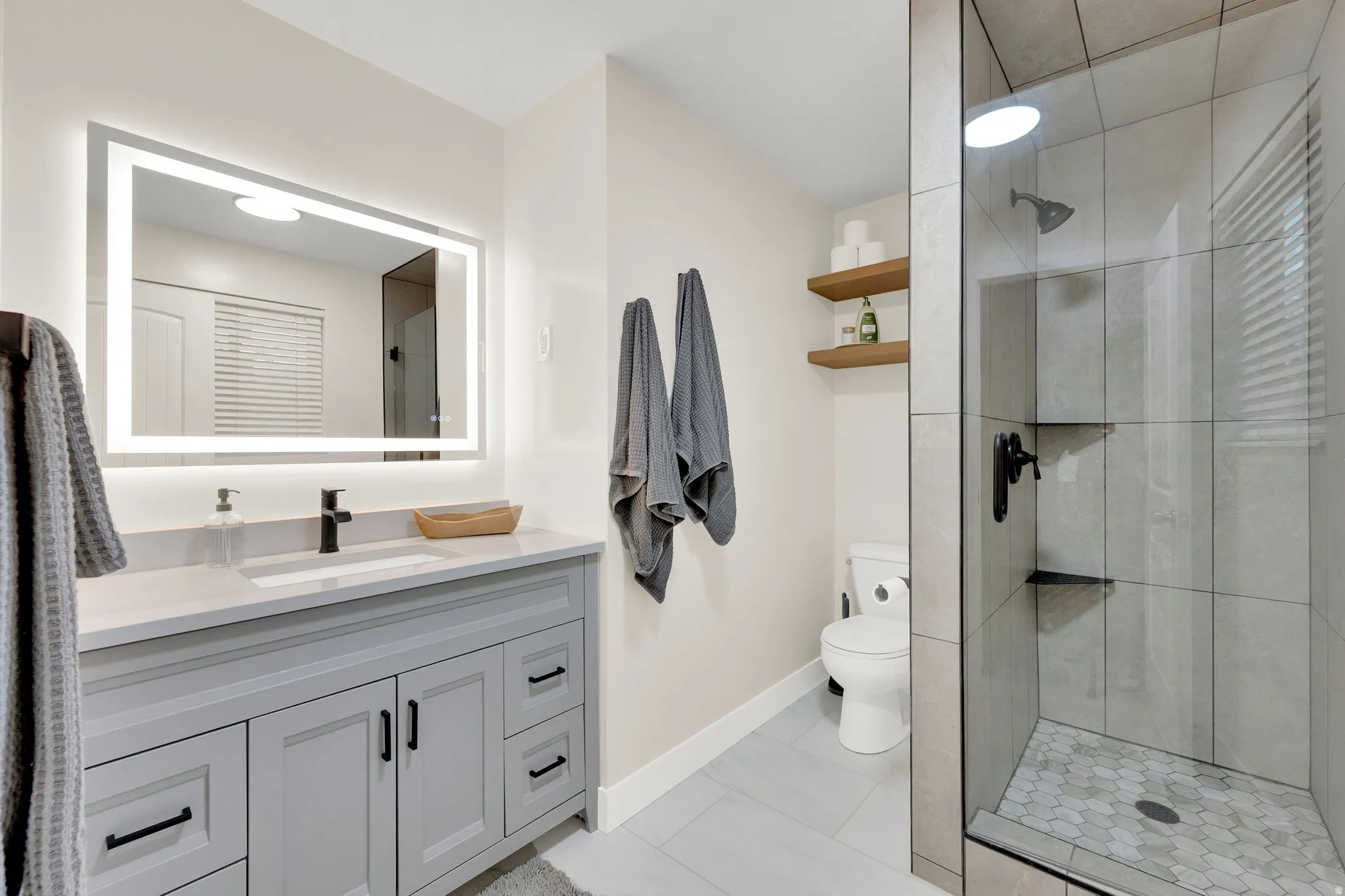 Modern bathroom featuring a glass-enclosed shower with a hexagon tile floor, a light gray vanity with a single sink, a lighted mirror, and matte black hardware
