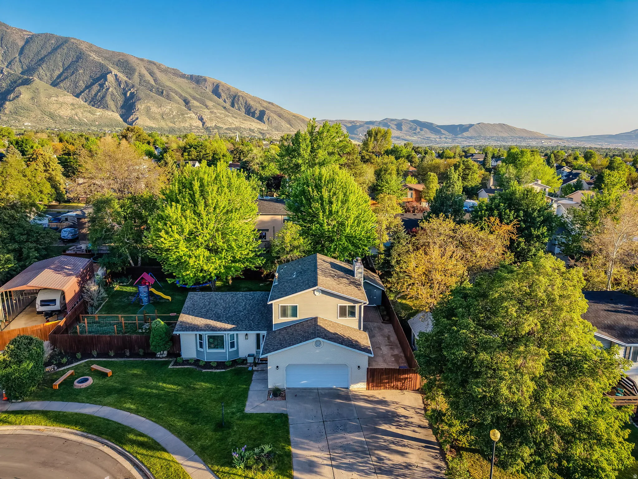 Two-story residence featuring a two-car garage, mature trees, and a spacious front lawn with a built-in fire pit