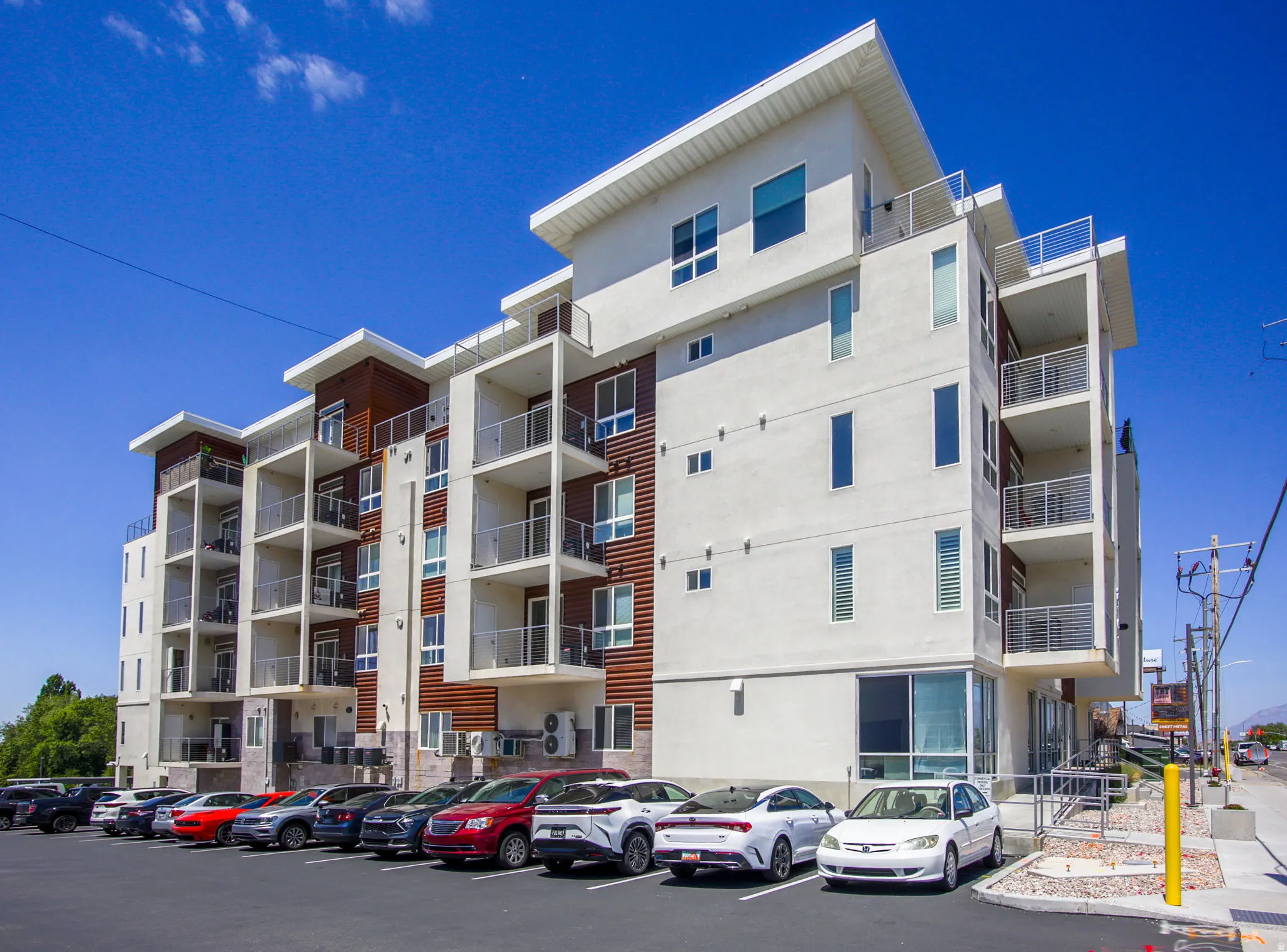Modern multi-story building featuring a stucco exterior, wood panel accents, multiple private balconies with metal railings, and a ground-level parking area
