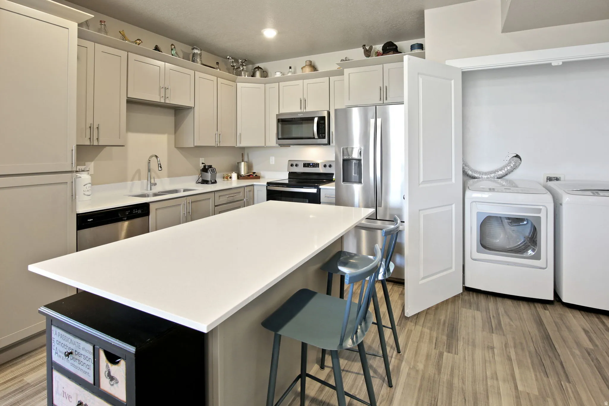 Kitchen featuring an island with white countertop, stainless steel appliances, light-toned cabinetry, and wood-finish flooring