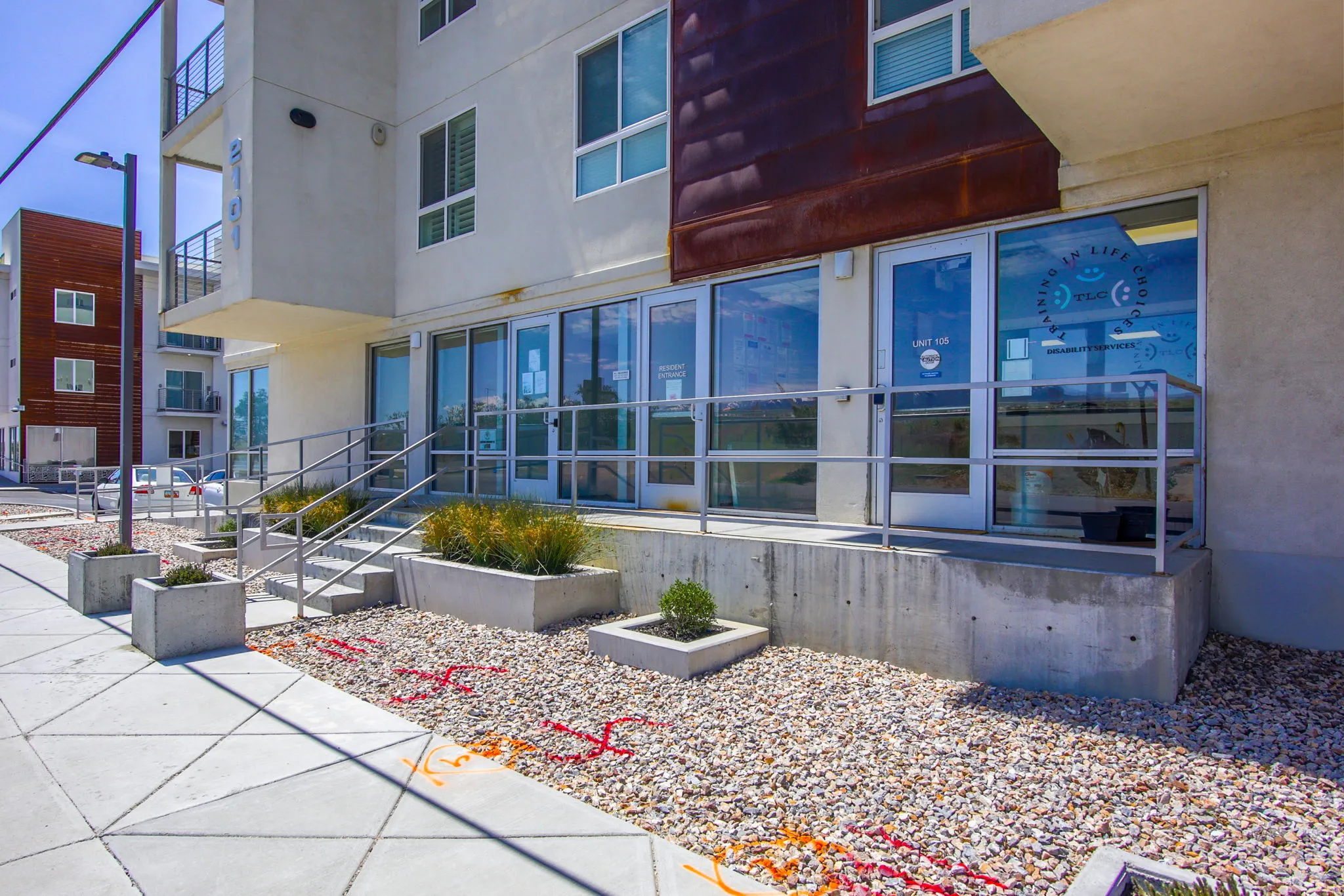 Modern building exterior featuring a textured brown facade accent, concrete planter boxes with established greenery, a concrete walkway, and large storefront windows with silver framing