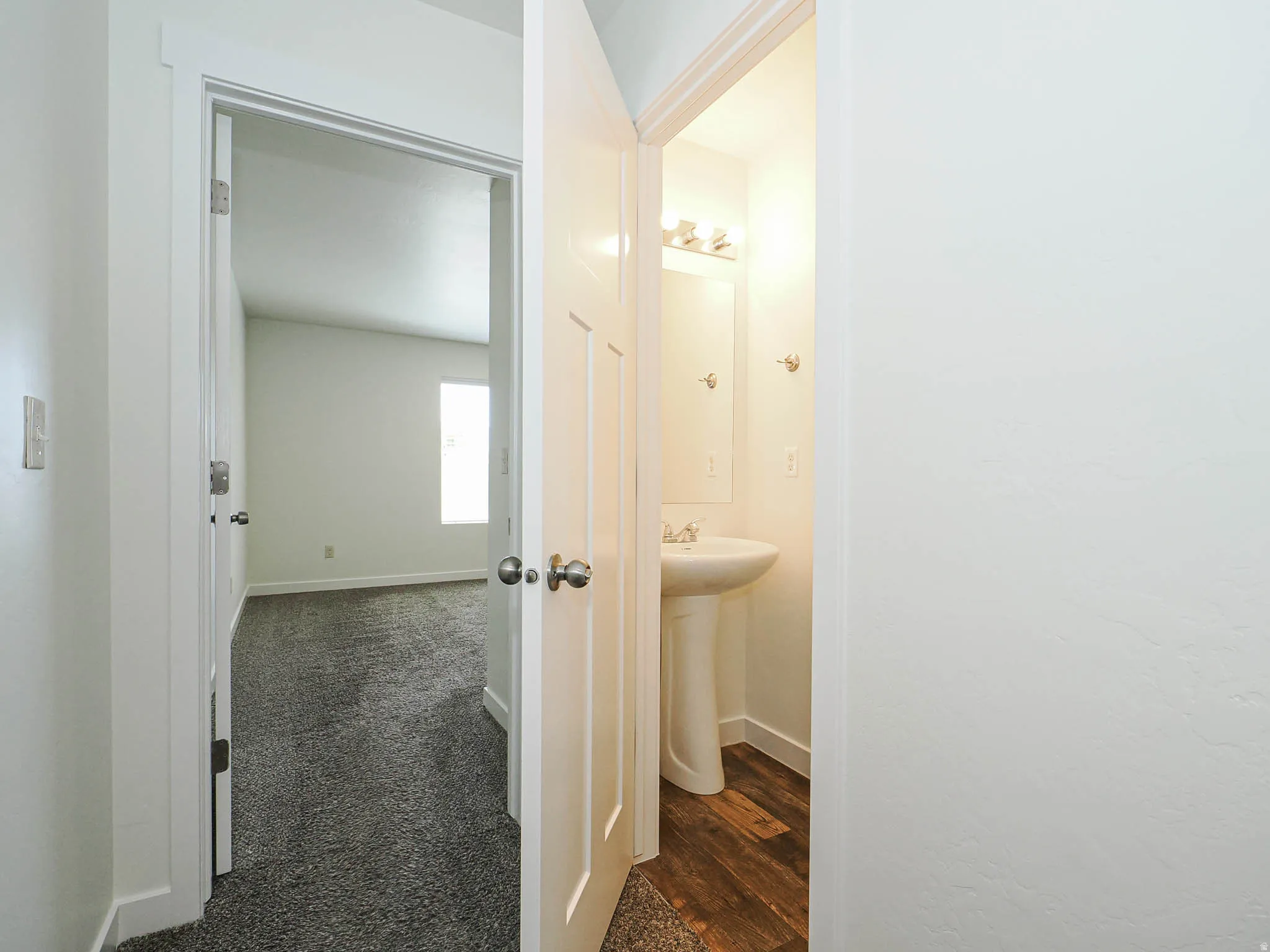 Interior hallway featuring crisp white walls and doors, leading to a carpeted room with natural light and a bathroom with wood-finish flooring and a pedestal sink