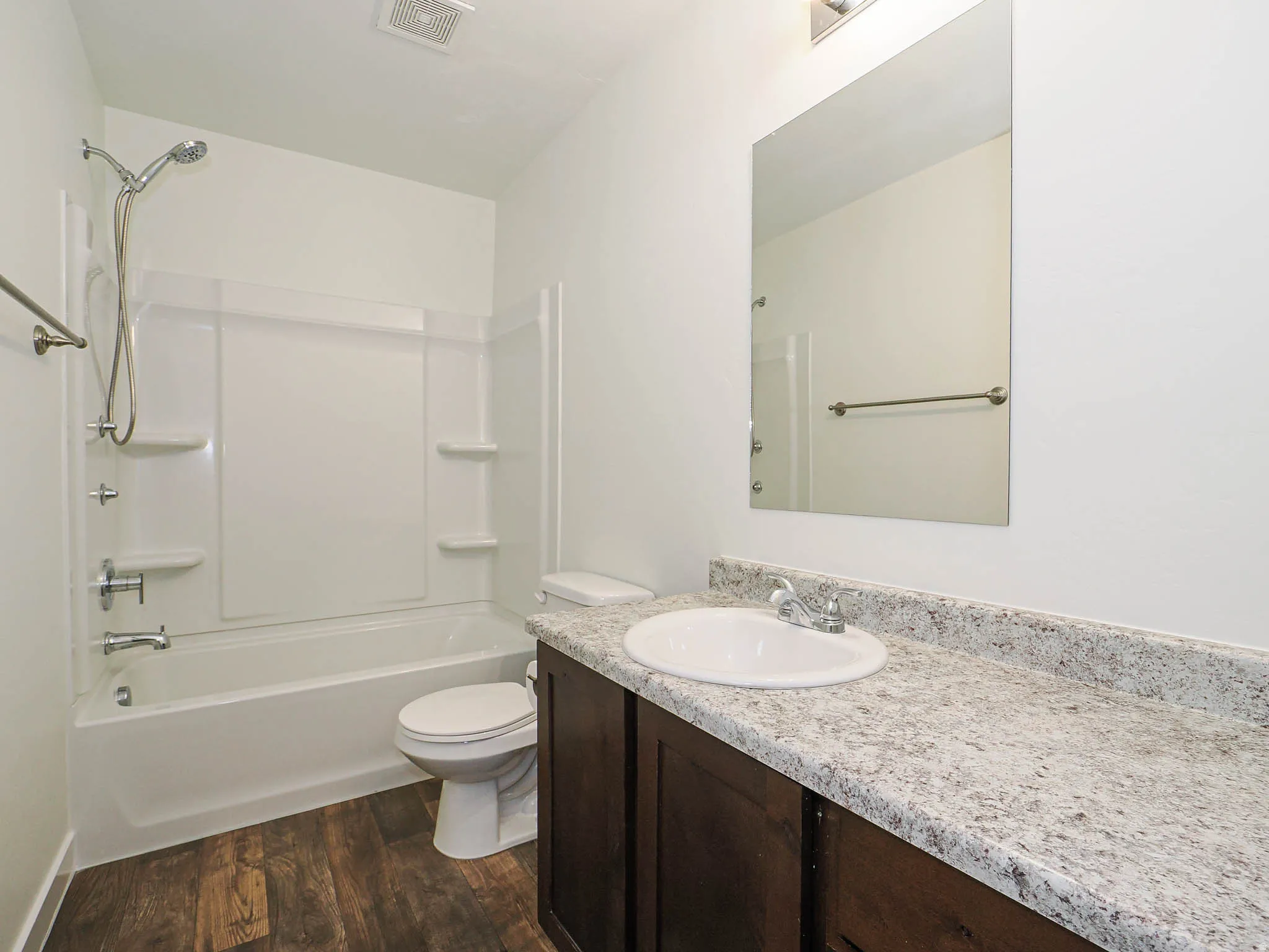 Bathroom featuring a white bathtub and shower surround, a dark wood-finish vanity with a speckled countertop, a large wall-mounted mirror, and wood-finish flooring