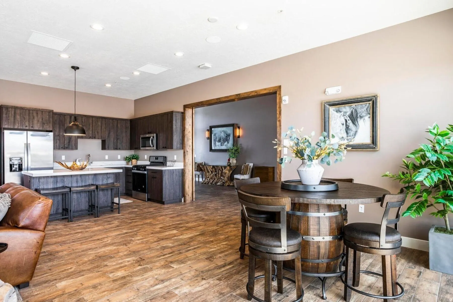 Open-concept interior featuring wood-finish flooring, a kitchen with dark cabinetry and stainless steel appliances, a kitchen island with seating, recessed lighting, and a wide entryway with wood trim