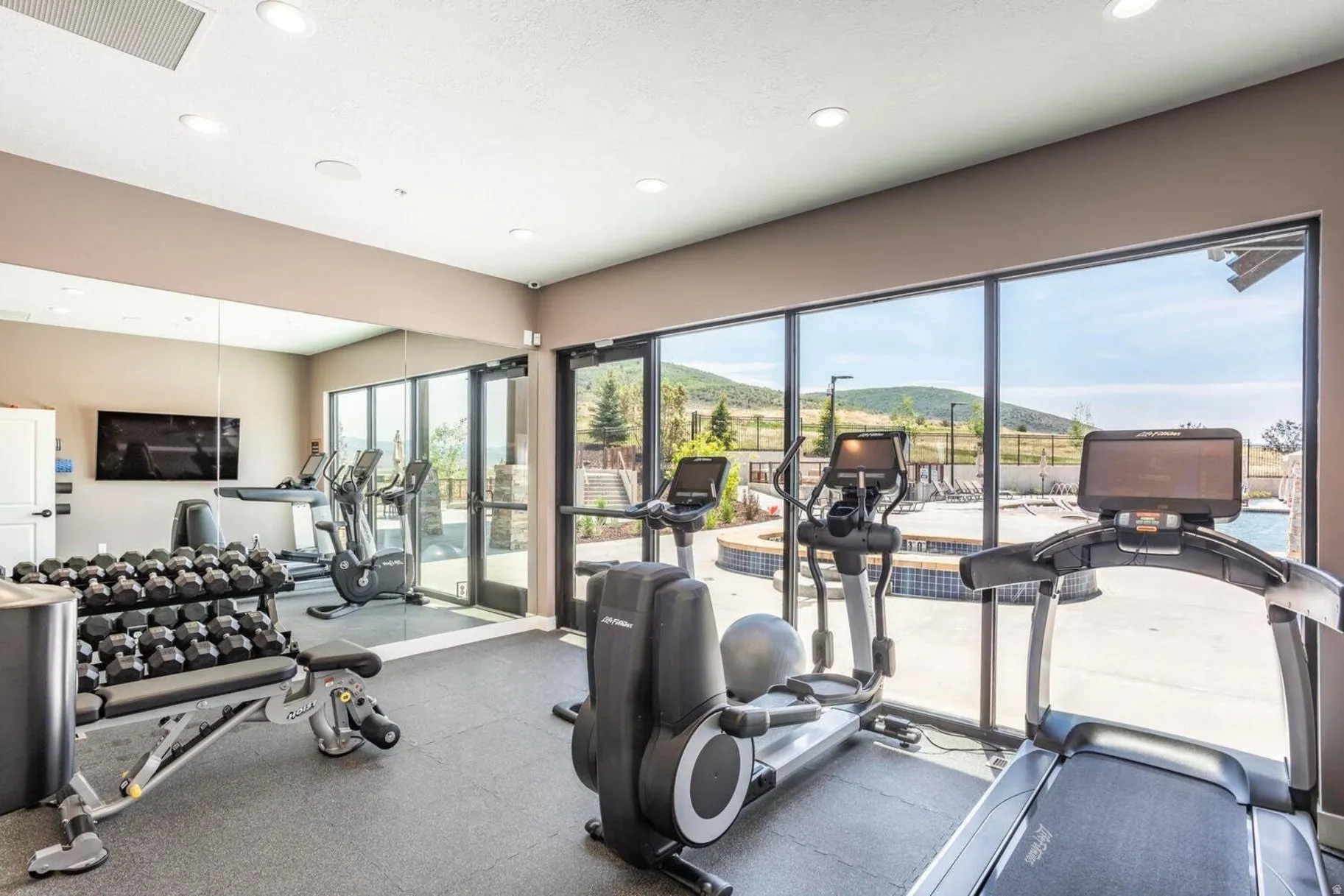 Spacious fitness room featuring expansive floor-to-ceiling windows, recessed lighting, wall-mounted mirrors, a mounted television, and a durable rubberized floor