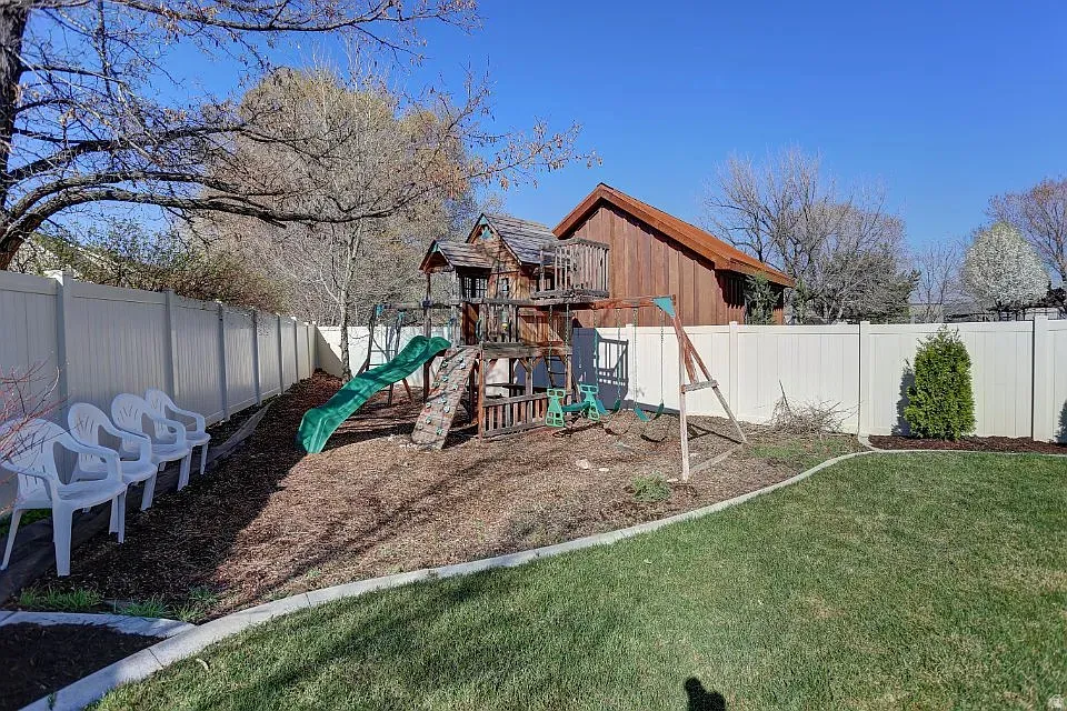 Spacious backyard featuring a manicured lawn, a white privacy fence, and a multi-level playset with slides and swings