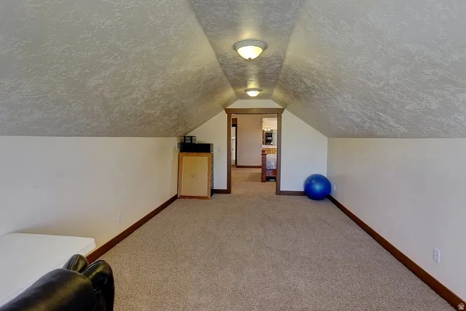 Carpeted upper-level bonus room featuring a vaulted ceiling with textured finish, recessed lighting, and a doorway leading to additional finished space