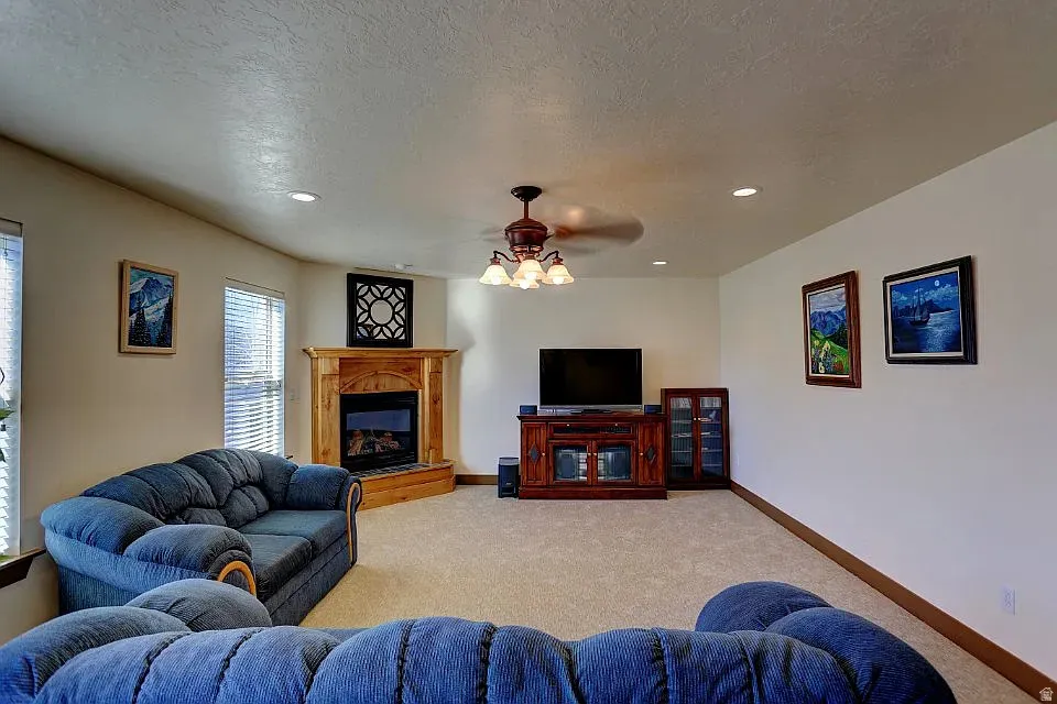 Carpeted living space featuring a corner fireplace with wood mantel, recessed lighting, and a ceiling fan with integrated light fixture