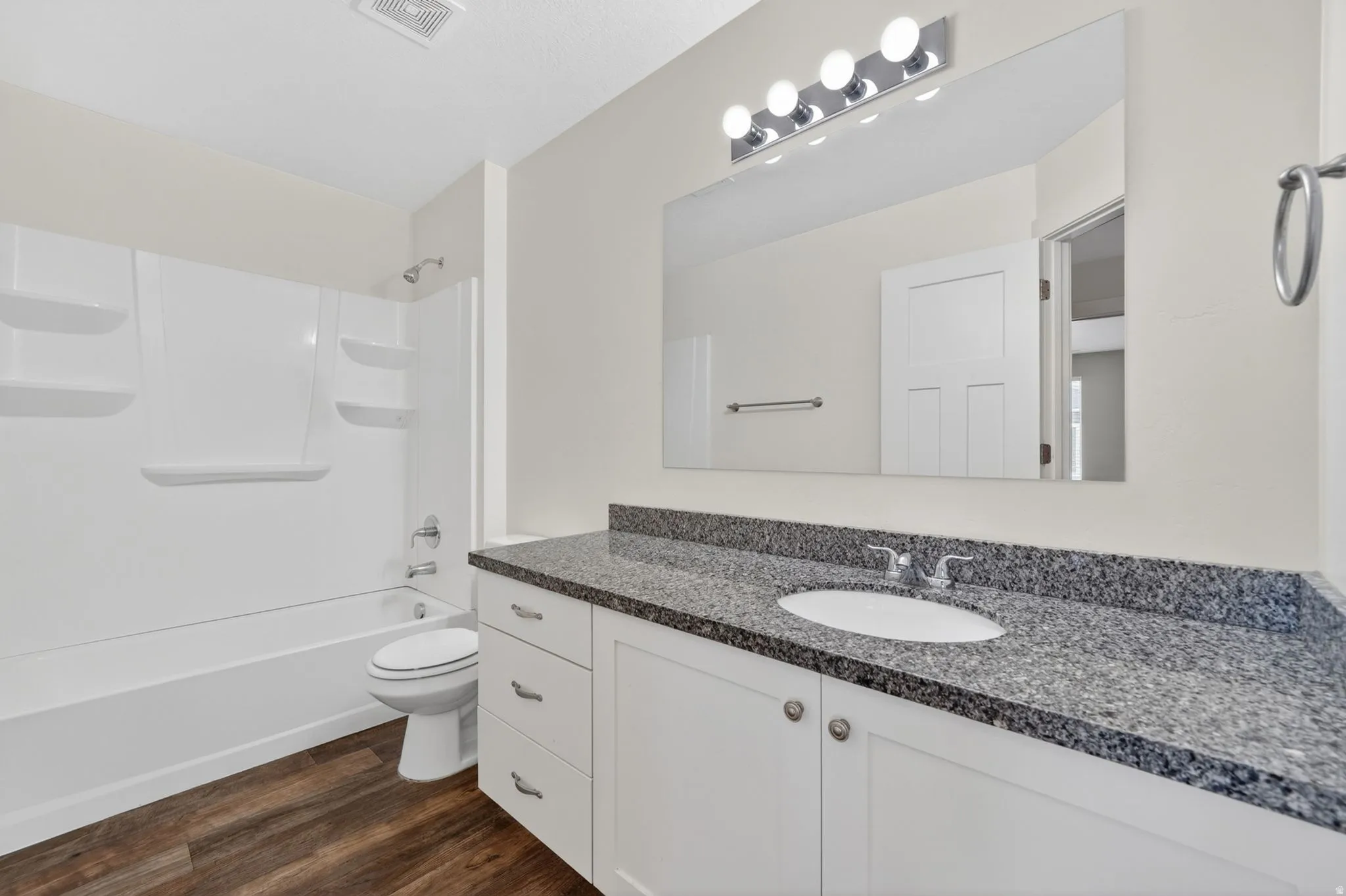 Bathroom featuring wood-finish flooring, a white vanity with brushed nickel hardware, a dark speckled countertop, an undermount sink, and a white shower-over-tub combination with built-in shelving