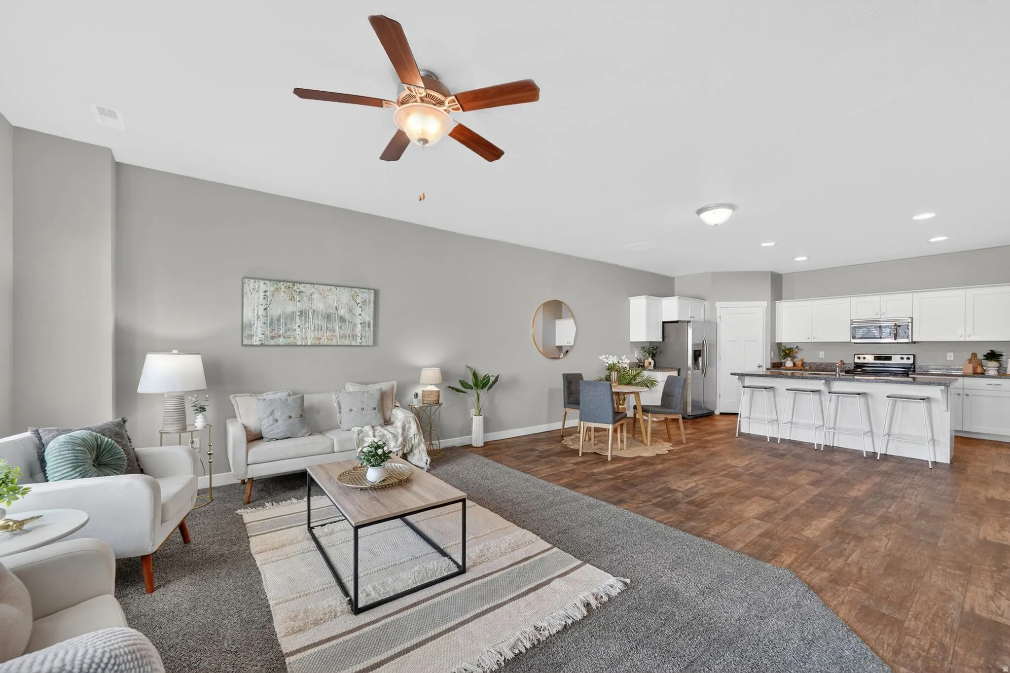 Open-concept living space featuring a living area with a ceiling fan, wood-finish flooring in the dining area, and a kitchen with white cabinetry, stainless steel appliances, and an island with bar seating