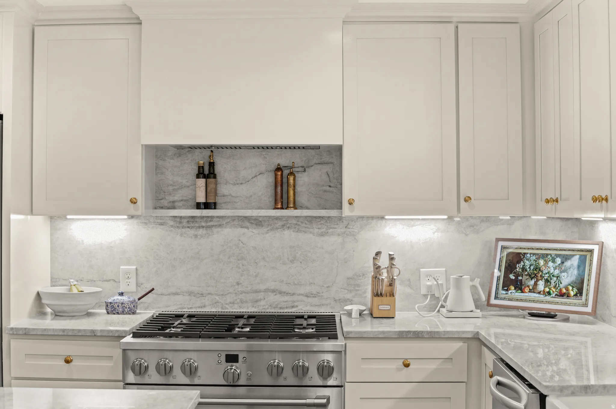 Gourmet kitchen featuring a stainless steel range, light-toned cabinetry with gold-toned hardware, stone countertops, a full-height stone backsplash, and recessed under-cabinet lighting