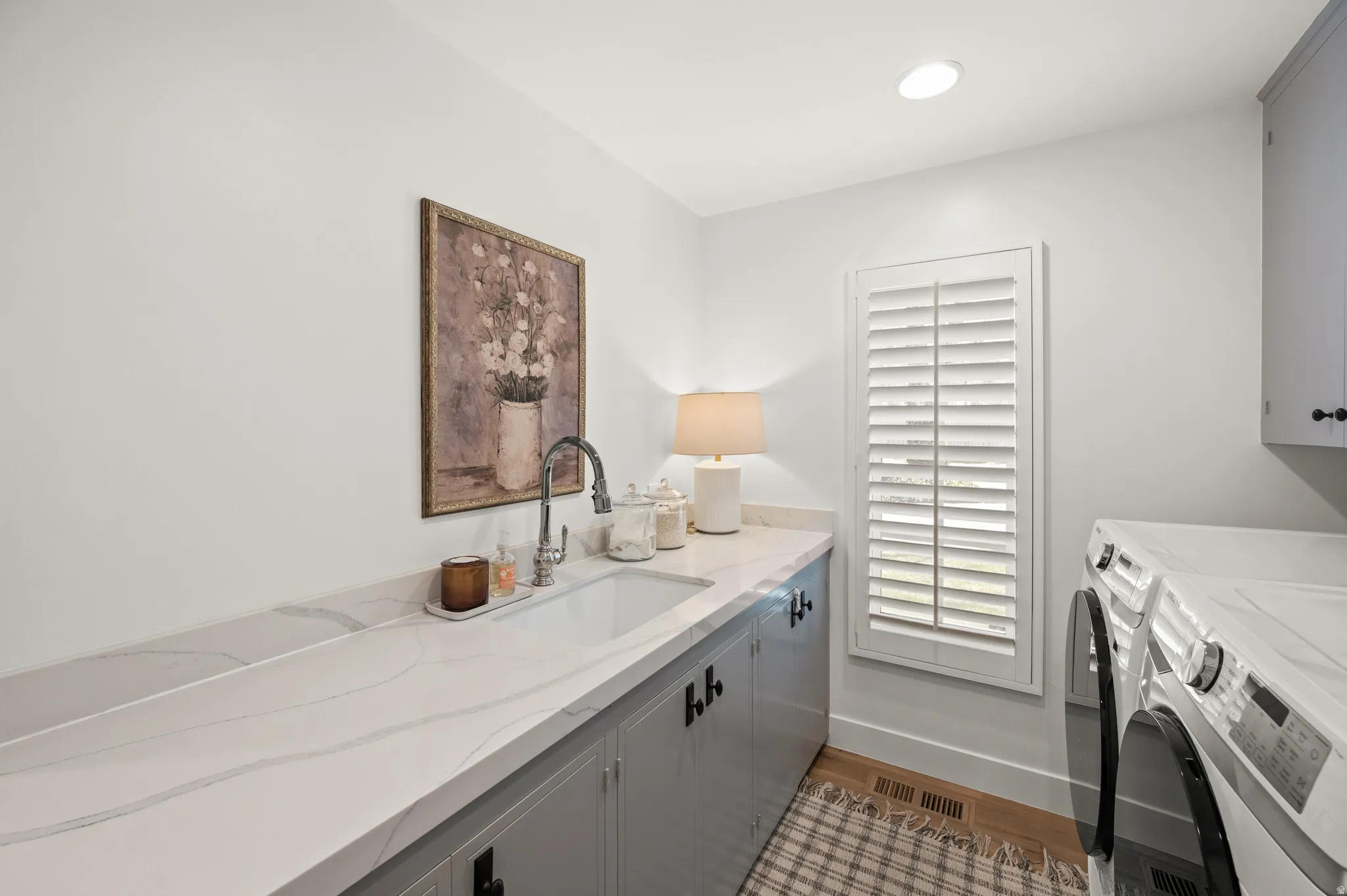 Utility room featuring a built-in sink, stone-finish countertop, shaker-style cabinetry with matte black hardware, and window with plantation shutters