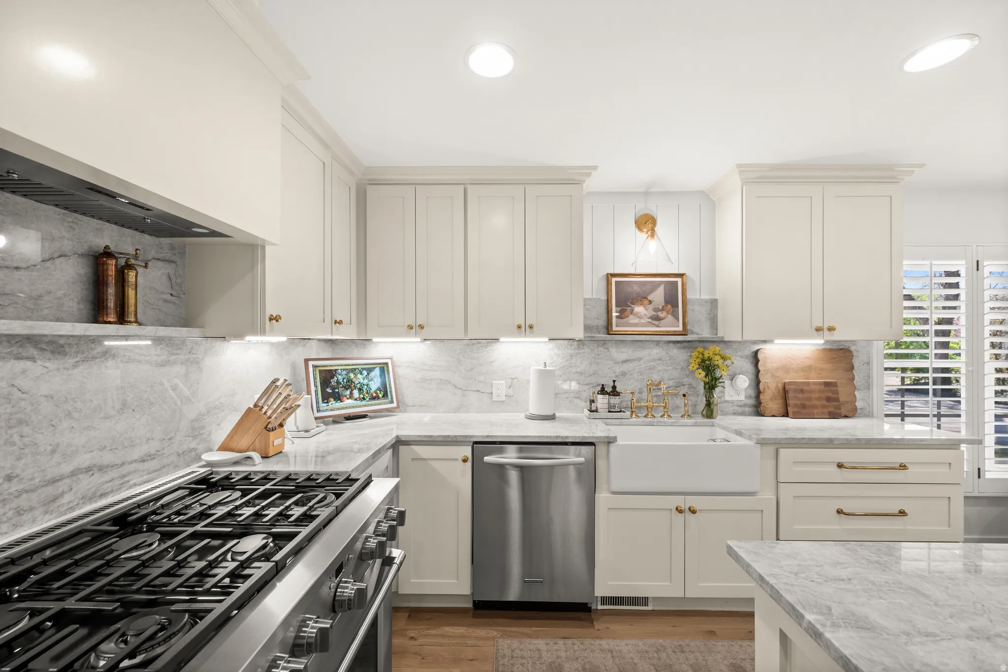 Kitchen featuring a stainless steel range with a built-in hood, light-toned cabinetry, stone countertops with a matching backsplash, a farmhouse sink, and plantation shutters